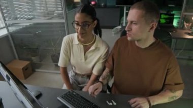 Orta boy beyaz erkek programcı. Dövmeli kolları var. Bilgisayar masasında oturuyor ve melez kız arkadaşına ders veriyor.