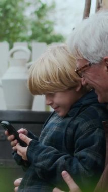 Dikey yan görüntü, mutlu büyükbaba ve torunun akıllı telefona bakıp verandada bahçeli güzel bir kır evinin verandasında otururken kahkahalarını paylaştığı orta boy bir fotoğraf.