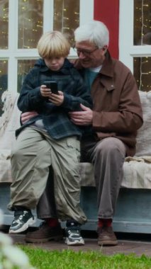 Gülümseyen beyaz saçlı büyükbaba ve küçük torununun rahat verandada birlikte oturup, çiçeklerle çevrili akıllı telefona ve sıcak aydınlatmaya baktıkları hassas anı paylaşmaları.