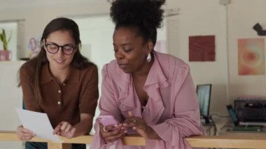 İki farklı kadın meslektaşın fikir alışverişinde bulunduğu orta boy bir fotoğraf, biri telefonu tutarken diğeri de rahat ofis ortamının ortak çalışma ortamında belgeyi gözden geçirirken.