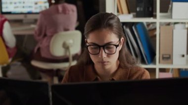 Modern ofis ortamında çift monitörde çalışan gözlüklü ve kahverengi polo süveter giyen odaklanmış beyaz bir kadının göğsü. Takım arkadaşları bulanık arka planda iş birliği yapıyorlar.