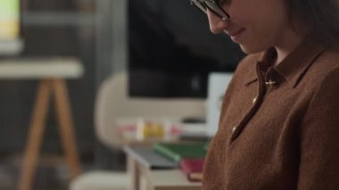 Gözlüklü ve kahverengi polo kazaklı gülümseyen genç beyaz bir kadının iş günü bulanık ofis zemininde otururken telefonuyla etkileşim kurduğu bir kare.