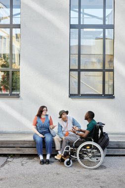 Engelli genç Afrikalı Amerikalı 'ya dikey bakış açısı açık. Açık havada arkadaşlarıyla sohbet ediyor. Beyaz bina duvarına yaslanmış, güneş ışığı fotokopi alanı var.