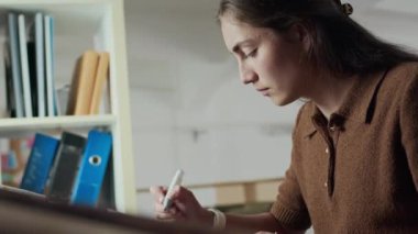 Yan görünüm: Ofis masasında kağıt üzerinde resim çizerken kahverengi polo kazaklı, mavi fosforlu kalem kullanan beyaz bir kadın.