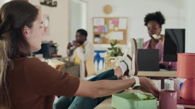 Kafkasyalı genç bir kadın, rahat ofis ortamında yemek yerken ve iki Afro-Amerikan takım arkadaşıyla sohbet ederken ayakları masanın üzerinde oturuyor.