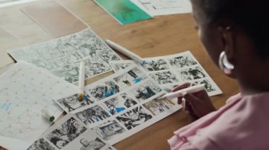 Afrika kökenli Amerikalı kadın sanatçı, görsel efektleri artırmak için renkli işaretler kullanarak, ahşap çalışma istasyonunda detaylı bir taslak üzerinde çalışıyor.