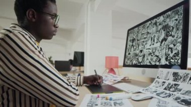 Ekranda taslağı olan bilgisayara bakarken, modern ofis ortamında masada otururken grafik tabletinde çizim yapan genç siyahi erkek ressamın belini kaldırın.