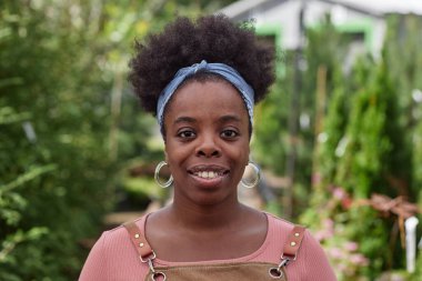 Yeşil bahçe çiftliğinde çalışırken kameraya gülümseyen Afrikalı Amerikalı genç bir kadının ön manzara portresi.