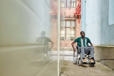 Engelli Afrikalı Amerikalı adamın geniş açılı görüntüsü. Şehirde tekerlekli sandalye kullanıyor ve binalar arasındaki dar kaldırımda hareket ediyor.