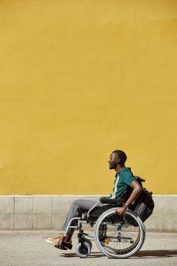 Engelli Afrikalı Amerikalı 'nın dikey yan görüntüsü. Şehir dışında tekerlekli sandalye kullanıyor. Sarı renkli duvar fotokopi alanı karşılıyor.