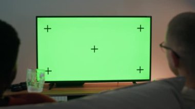 Akşam vakti rahat bir dairede, yeşil ekran TV karşısında sohbet eden iki adamın arka plan görüntüsü bira ve atıştırmalıklarla rahat bir kanepede otururken.