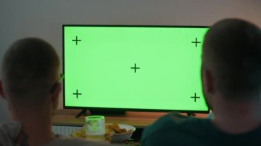 Arka planda iki erkek arkadaşın modern oturma odasındaki yeşil ekran TV ekranında oturup sohbet ederken ve abur cubur ve bira içerken görüntüsü var.