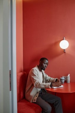 Kafe masasında renkli minimalist tasarım ile otururken not defteri planlama programı yazan konsantre Afrikalı Amerikalı iş kadınının dikey görüntüsü, kopyalama alanı