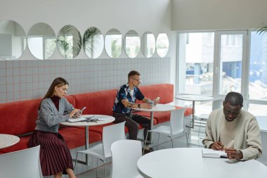 Afrika kökenli Amerikalı iş adamının çalışma alanındaki masada otururken deftere yazdığı geniş açı çekimi. Modern minimalist tasarıma sahip rahat bir kafede çalışan iş adamları