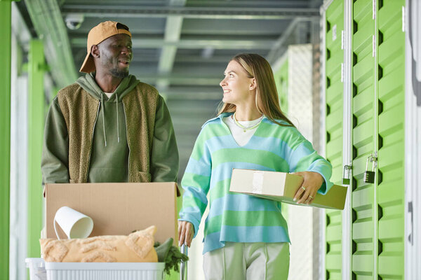 Medium shot of cheerful African American man pushing dolly cart chatting with girlfriend moving cardboard boxes into self storage unit during apartment decluttering