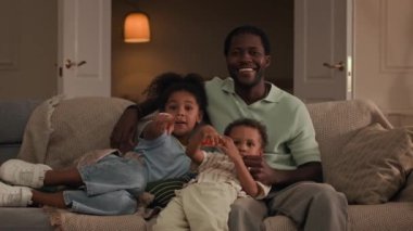 Orta boy bir Afrikalı Amerikalı baba, yeni yürümeye başlayan oğlu ve küçük kızı kanepede oturup birbirlerine sarılırken ve rahat, modern oturma odasında televizyon izlerken.