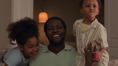 Genç Afrikalı Amerikalı babayı sevmenin orta boy portresi, küçük oğlu ve küçük kızı birbirine sarılıyor, gülümsüyor ve evdeki güzel modern oturma odasında kameraya bakıyor.