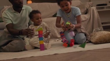 Küçük Afrikalı Amerikalı kardeşler, babasıyla koltuğun yanındaki plastik bloklardan oyuncak kulelerle oynuyorlar, rahat modern oturma odasında birlikte gülüyorlar.