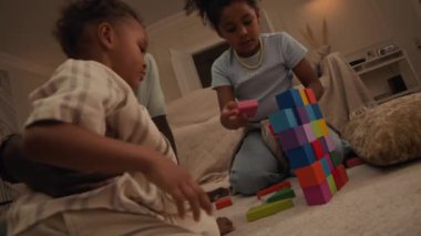 Küçük melez kızın oyuncak duvarını inşa ederken, rahat modern oturma odasındaki kanepenin yanındaki plastik bloklardan, siyah baba ve küçük kardeşin ona yardım ettiği düşük açılı bir kare.