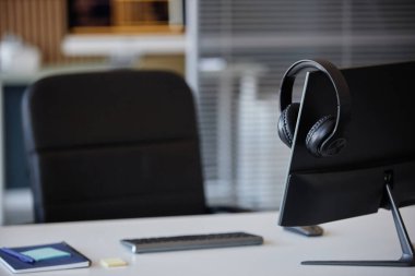 Ofis masasında kulaklık ve klavyeli bilgisayar monitörü var. Ofis sandalyesi ve raflar bulanık arkaplanda profesyonel çalışma alanı ayarları oluşturuyor
