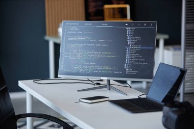 Monitörde bir kodlama arayüzüne sahip modern ofis, bir dizüstü bilgisayar ve akıllı telefon eşliğinde, çalışma alanında çeşitli teknoloji araçları ve cihazlar sergiliyor.