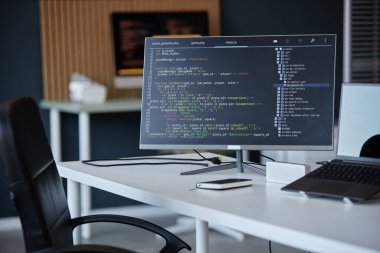 Yazılım kodlarını ve çeşitli masa ögelerini gösteren bir bilgisayar monitörünün yer aldığı modern ofis çalışma alanının resmi. Arkaplan, sandalye ve rafların görüldüğü bulanık ofis kurulumunu içeriyor
