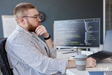 Modern ofiste çalışırken bilgisayar ekranına odaklanan sakallı bir adam. Kodlama monitöründe, kahve fincanı yanında.