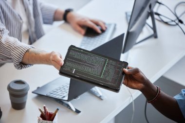 İş arkadaşları, modern bir ofis ortamında tablet kullanarak önemli bilgileri paylaşıyor ve arka planda bilgisayar çalışma istasyonları ve kahve bulunuyor.