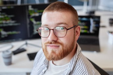 Modern ofis ortamında kod gösteren bilgisayar ekranlarıyla çevrili gözlüklü sakallı bir adamın portresi.
