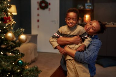 Yetişkin Afro-Amerikan anne portresi oğlunu kucaklıyor ve Noel ağacıyla şakalaşıyor. Tatilleri birlikte kutluyorlar.