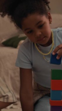 Siyahi babası ve küçük kardeşiyle birlikte rahat modern oturma odasında oyun oynarken koltuğun yanındaki plastik bloklardan oyuncak kulesi inşa eden melez bir kızın dikey görüntüsü.