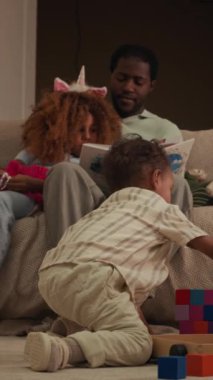 Babası ve ablasıyla yerde oynayan, pembe tek boynuzlu at saç bandı takmış gri kanepe üzerinde oturmuş modern oturma odasında kitap okuyan Afrikalı Amerikalı bir çocuğun dikey görüntüsü.