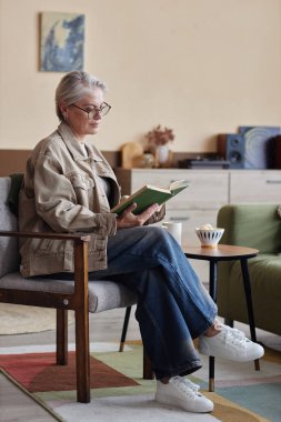 Rahat bir evde, sandalyede oturmuş, rahat bir şekilde kitap okuyan zarif, olgun bir kadının dikey portresi.
