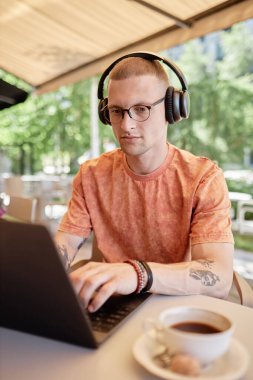 Kablosuz kulaklıklarla konsantre olmuş işadamının dikey görüntüsü klavye üzerinde çalışmak için dizüstü bilgisayar kullanırken kafenin açık oturma alanında çalışırken, boşluğu kopyalayarak.