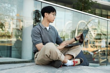 Orta büyüklükte Asyalı erkek bilgisayar programcısının dokunmatik ekranı izlerken kaldırımda cam inşaatının yanında otururken şehir caddesinde halka açık alanda çalışırken görüntüsü.