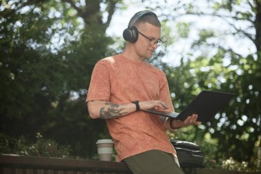 Kulaklıkla bilişim programcısının bilgisayarla kod yazarken görüntüsü. Müzikten keyif alırken doğa parkta bankta takılıyor. Açık havada çalışıyor, uzayı kopyalıyor.