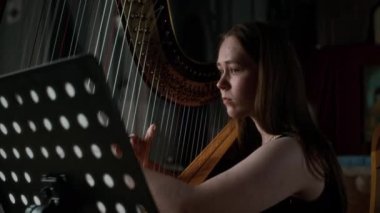 Kafkasyalı, zarif bir elbiseyle müzik standının önünde arp çalıp bulanık arka planda loş aydınlık katolik kilisesiyle skor arayan genç bir kadın arpçının orta boy yakın çekimi.