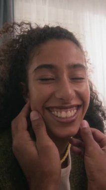 Uzun kıvırcık saçlı, ışıl ışıl gülümseyen, erkek arkadaşının nazikçe dokunduğu, sıcak, samimi bir ilişki kurduğu, rahat, hafif aydınlatılmış oturma odasındaki iki ırklı kadının dikey görüntüsü.