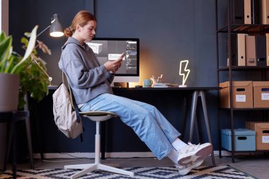 Masasında cep telefonu ekranına odaklanmış, rahat ofis ortamında günlük giysiler giyen genç bir kadın. Arkaplanda kutular, dekor ve saksı bitkisi olan raflar