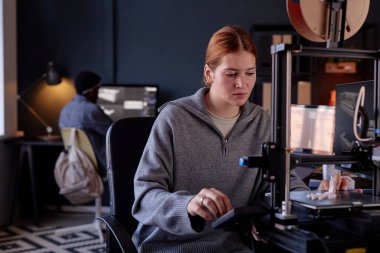 Meslektaşı arka planda yoğun ve teknolojik bir ortam gösteren ekrana odaklanırken bir kadın 3D yazıcıyla çalışıyor.