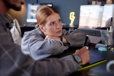 Genç bir kadın, modern ofis ortamında bir masada otururken kafasını ellerinin üzerinde dinlendirirken, düşünceli bir şekilde uzaklara bakıyor.