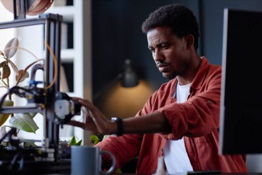 Adam 3D yazıcıyla karmaşık işler yapıyor. Konsantrasyon ve hassasiyeti vurguluyor. Çalışma alanı bitkileri ve yaratıcı bir ortamı vurgulayan çeşitli araçları içerir.