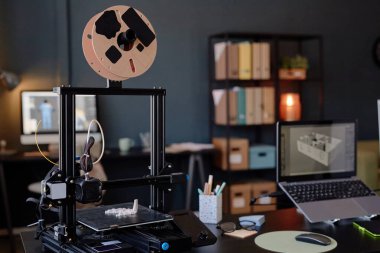 Masasında gelişmiş 3D baskı kurulumu olan modern atölyenin detaylı görüntüsü ve ekranda dizayn eden dizüstü bilgisayar. Arkaplanda kitap ve belgeli raflar yer alıyor