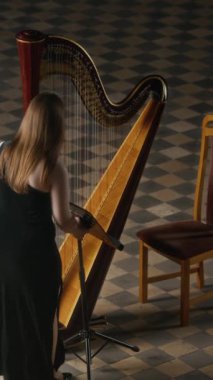 Siyah cüppeli, uzun, bol saçlı, müzik sehpası taşıyan, enstrümanın yanına oturan ve salonda dama tahtası zeminiyle gösteri yapmaya hazırlanan beyaz bir kadının dikey açılı çekimi.