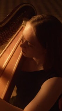 Dikey yüksek açılı, zarif, beyaz bir kadının klasik arp çalarkenki görüntüsü. İçerideki yumuşak, sıcak ışığın büyüleyici parıltısıyla aydınlatılmış.