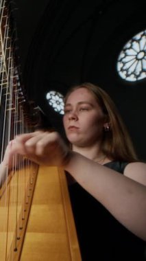 Dikey düşük açı orta boy beyaz kadın arpçının zarif vitray pencereler altında loş ışıklı katolik kilisede klasik arp çalarken görüntüsü.