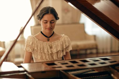 Portrait of young lady wearing lace dress with black choker necklace playing grand piano in softly lit classic room, copy space clipart