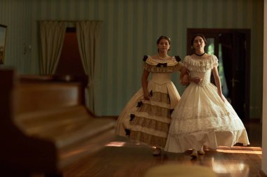 Full length portrait of two young ladies walking together towards camera in voluminous classic dresses across wooden floor in softly lit vintage ballroom, copy space clipart