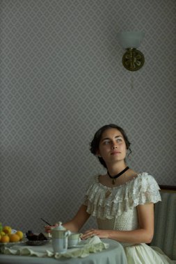 Klasik dantel elbiseli genç bayanın dikey portresi mektup yazıyor ve çay takımıyla zarif masada otururken dalgın dalgın yukarı bakıyor, boşluğu kopyalıyor.