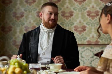 Portrait of bearded young gentleman smiling at young lady during elegant tea party in vintage setting clipart